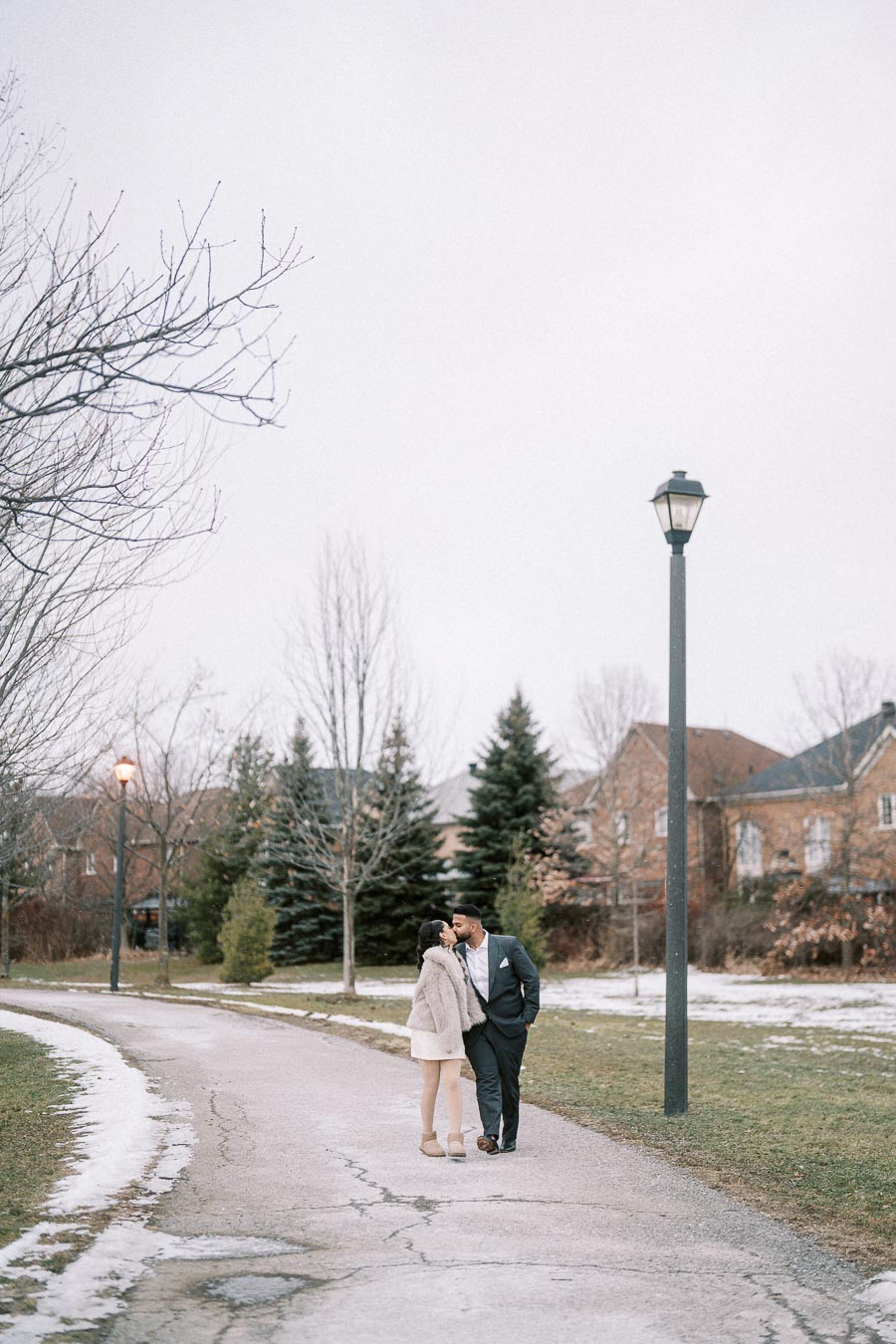 A couple kisses while walking down a snowy path in a suburban neighborhood, with bare trees and lamp posts lining the street on a cloudy winter day.