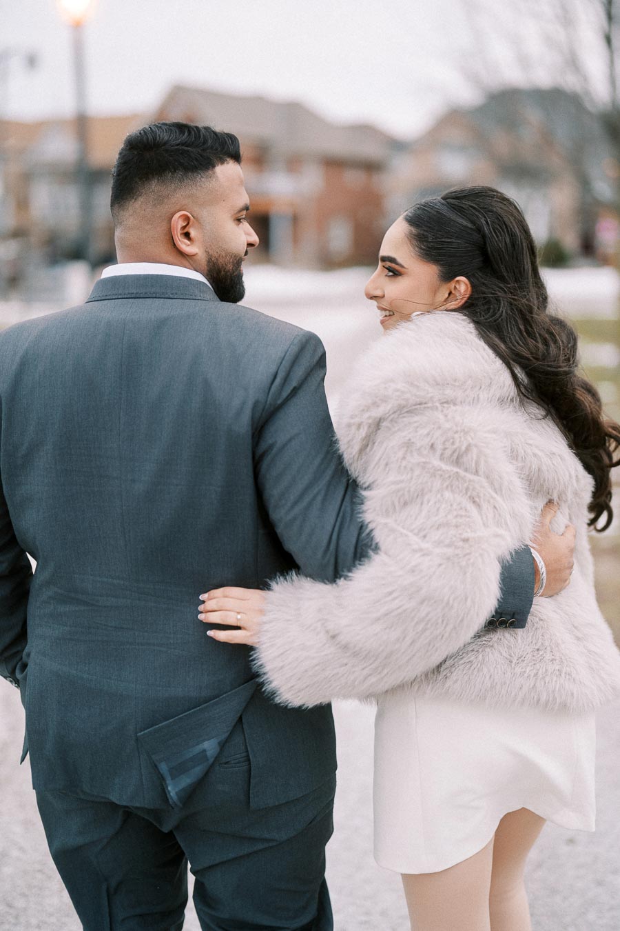 Couple walking together outdoors, dressed in elegant attire; man in a gray suit and woman in a white fur coat.
