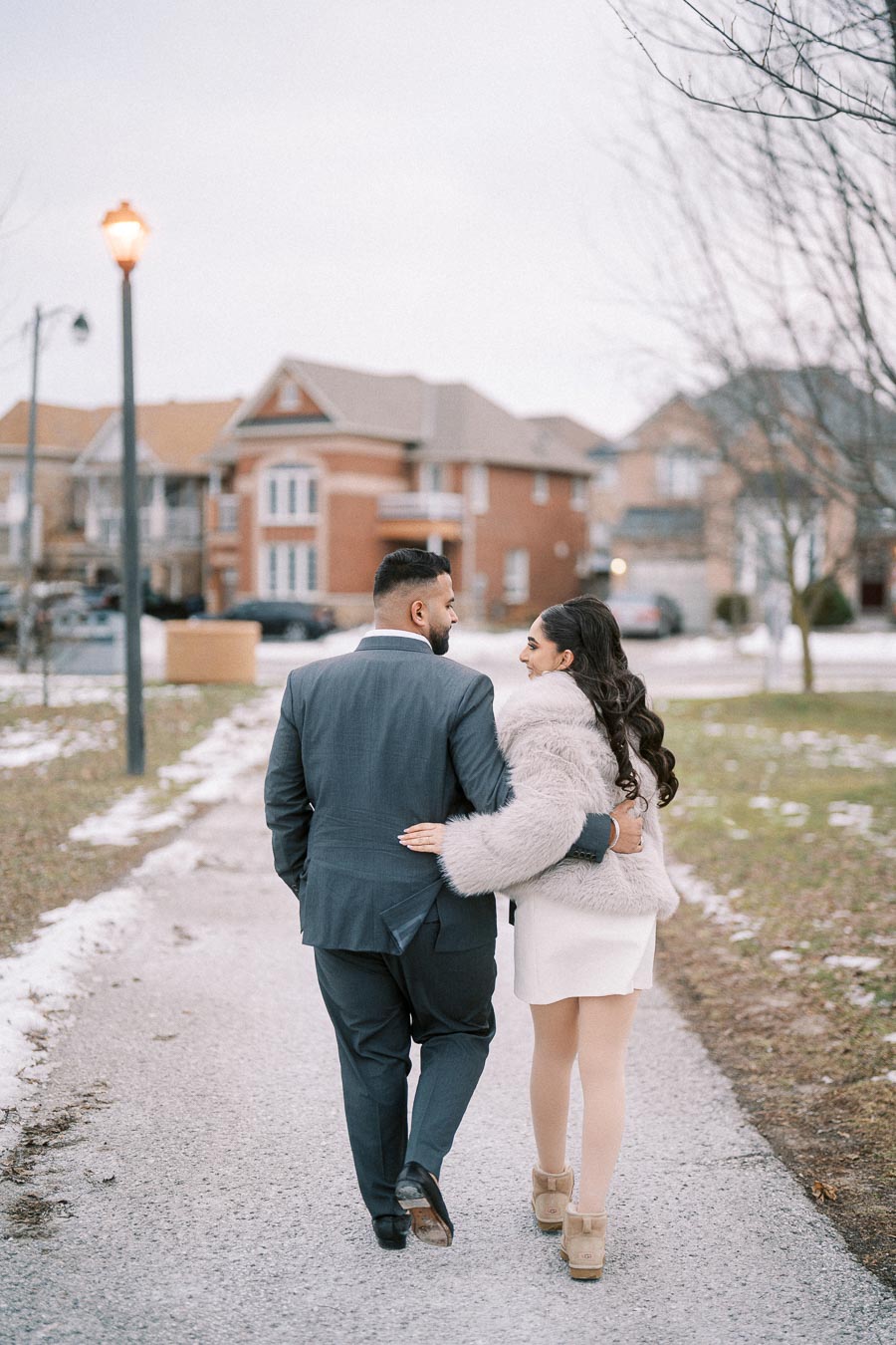 A couple walking arm in arm on a snowy path in a suburban neighborhood, dressed in formal attire with the woman wearing a fur coat and the man in a suit, surrounded by brick houses in the background.