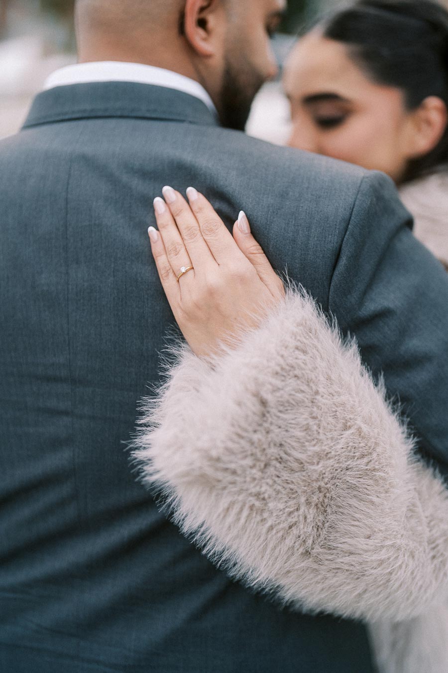 A woman in a furry coat embraces a man in a suit, showcasing her engagement ring with a delicate touch on his shoulder.