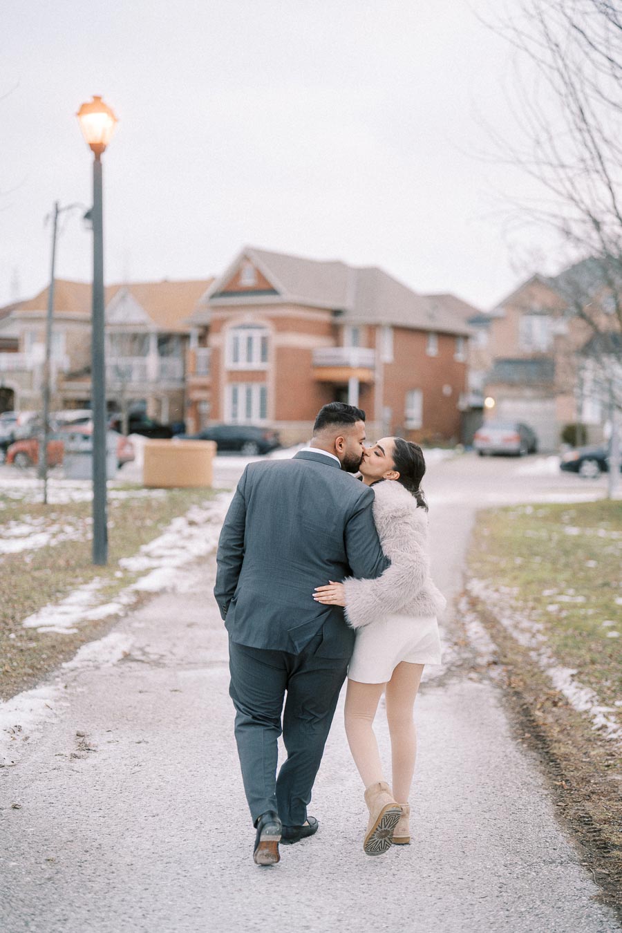 A couple walking down a snowy suburban street, dressed warmly, with the woman in a white outfit and fur coat and the man in a suit, sharing a kiss as they embrace.