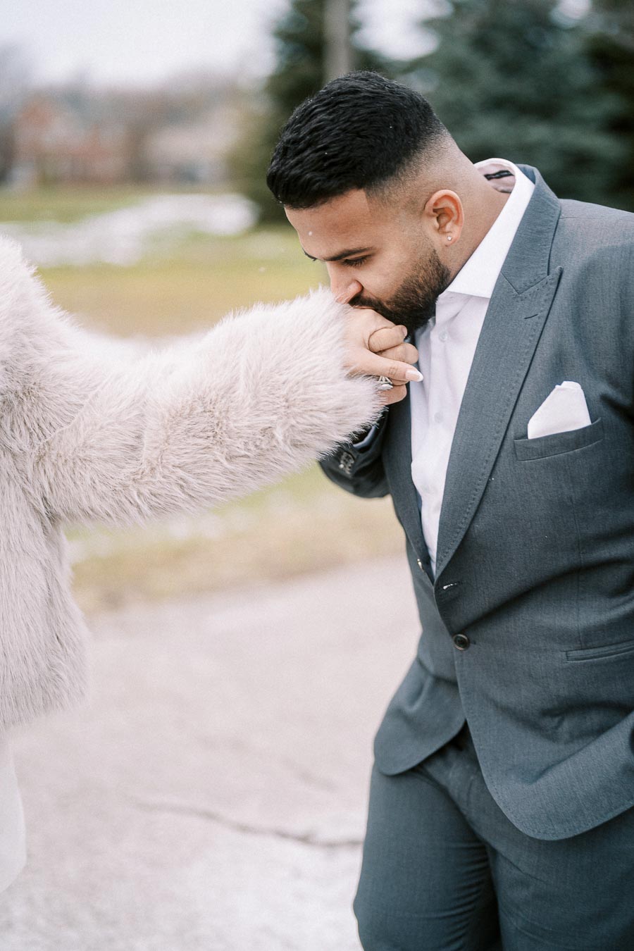 A man in a gray suit gently kisses the hand of a person wearing a fluffy white coat outdoors, showcasing a tender and romantic moment.
