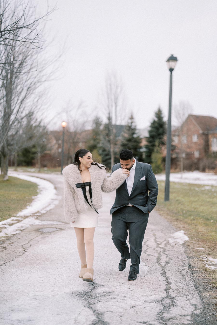 Couple walking along a snowy park pathway, woman in a fur coat and dress, man in a dark suit gently kissing her hand.