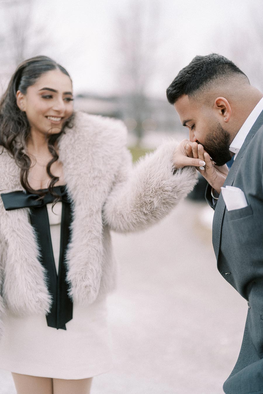Young woman in a stylish fur coat smiles as a man in a suit gently kisses her hand, creating a romantic moment outdoors.