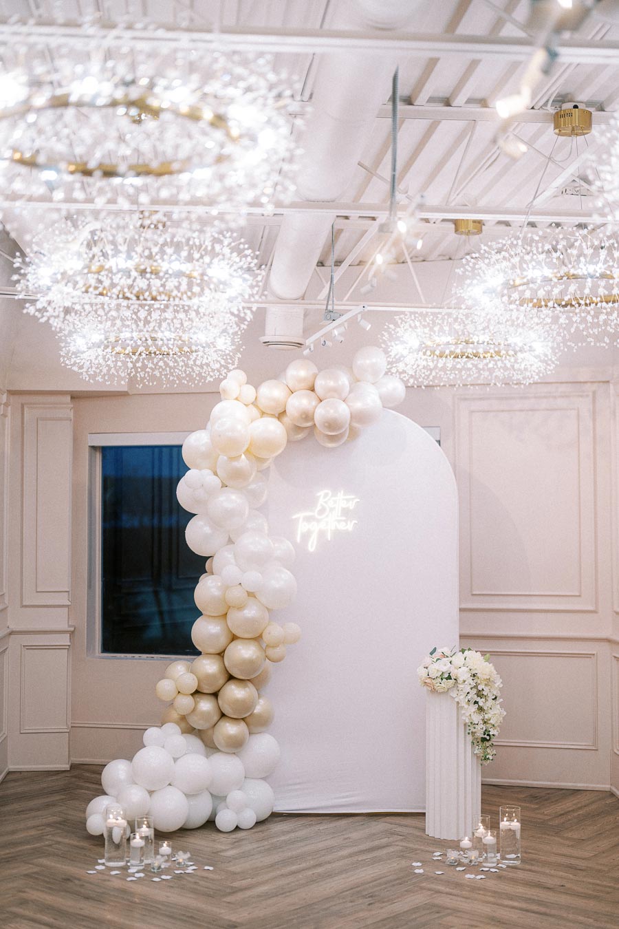 Elegant wedding venue with white and gold balloon arch, a neon sign reading Better Together, and decorative chandeliers. A floral arrangement on a pedestal and candles on the floor add to the sophisticated ambiance.