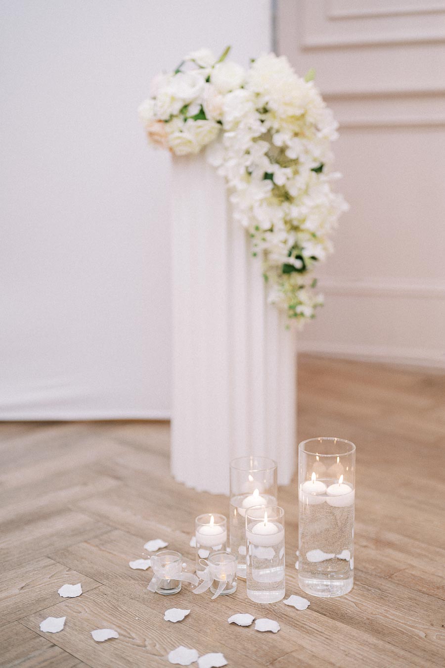 Elegant wedding decoration featuring white floral arrangement cascading over a tall pedestal, surrounded by floating candles in glass vases and scattered white rose petals on a wooden floor. Perfect for a sophisticated ceremony.