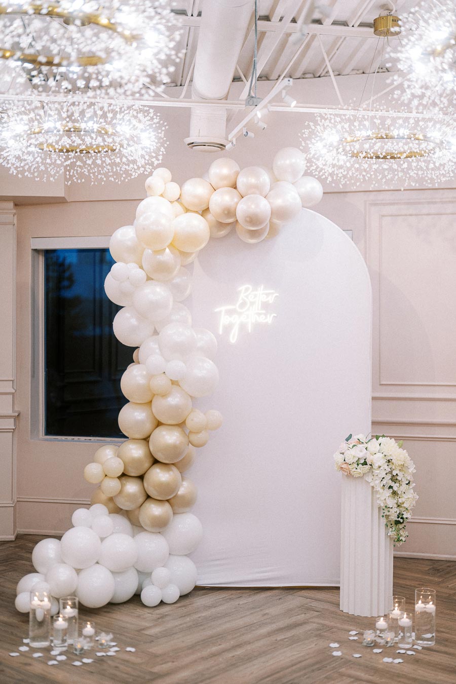 Elegant wedding backdrop featuring a white and gold balloon arch, with Better Together illuminated in neon. Decor includes floral arrangements on a white pedestal and candlelit ambiance on a wooden floor, under sparkling chandeliers.