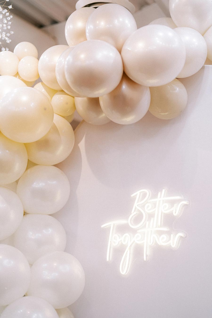 A decorative arrangement of creamy white balloons adjacent to a bright neon sign displaying the words Better Together against a soft beige background, creating an elegant and celebratory atmosphere.