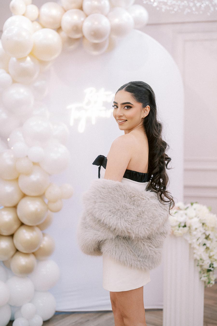 Young woman in elegant white dress and faux fur shawl at a sophisticated event with white and gold balloon decorations.