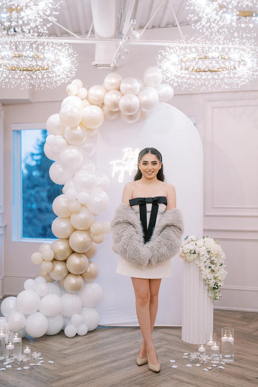 A woman in a cream dress and fur shawl stands in front of a stylish event backdrop featuring white and gold balloons, floral arrangements, and lit candles, creating an elegant and festive atmosphere.