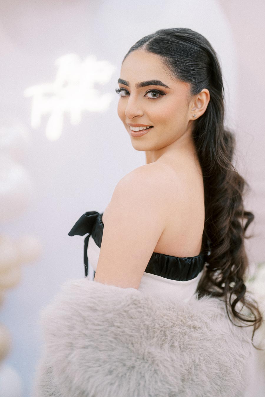 Alt text: Elegant woman with long dark hair and subtle makeup poses in a white dress with black accents and a fluffy gray shawl, showcasing a glamorous and sophisticated look.
