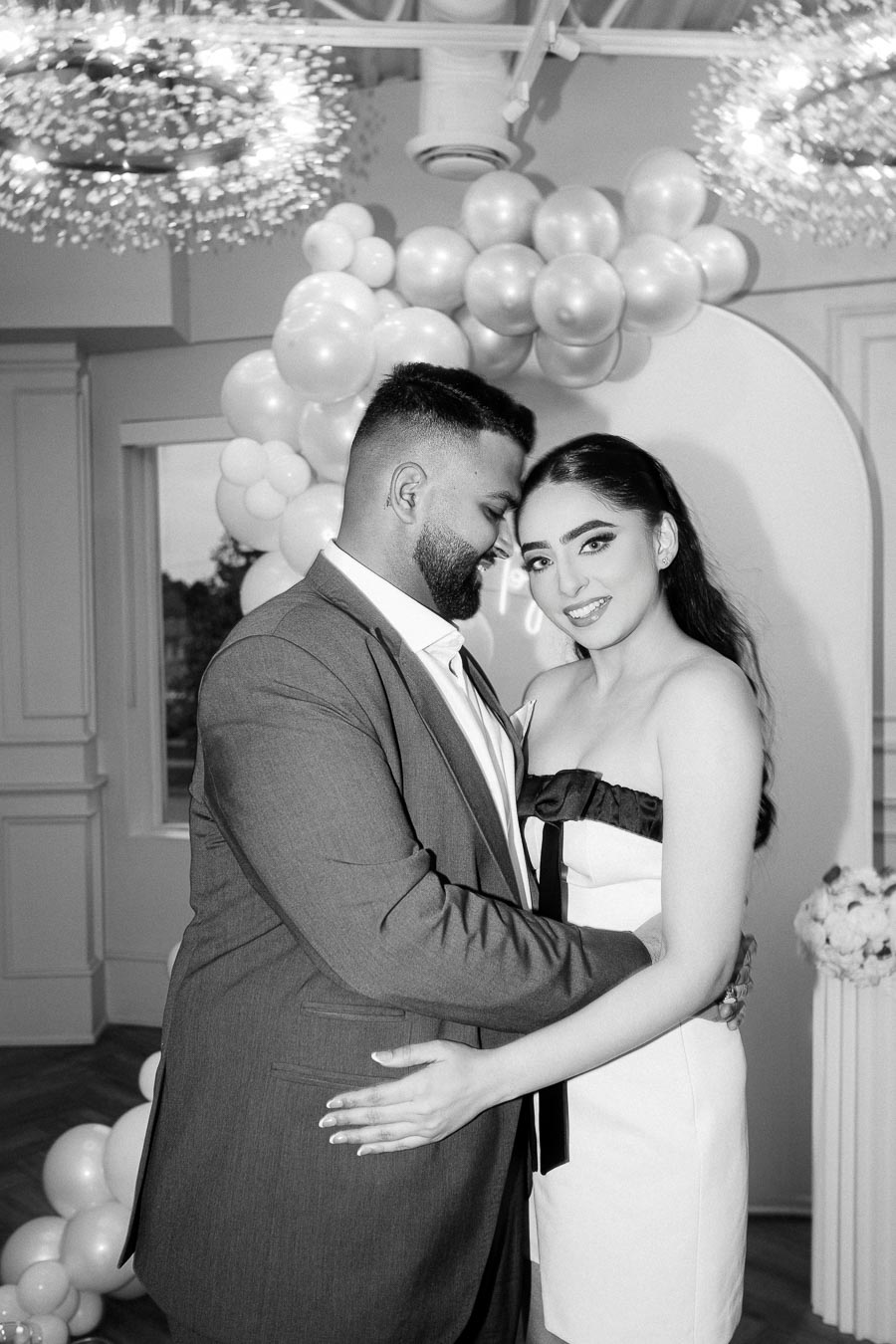 A couple embracing in a black and white photo against a backdrop of white balloons and elegant decor, showcasing a moment of joy and celebration.