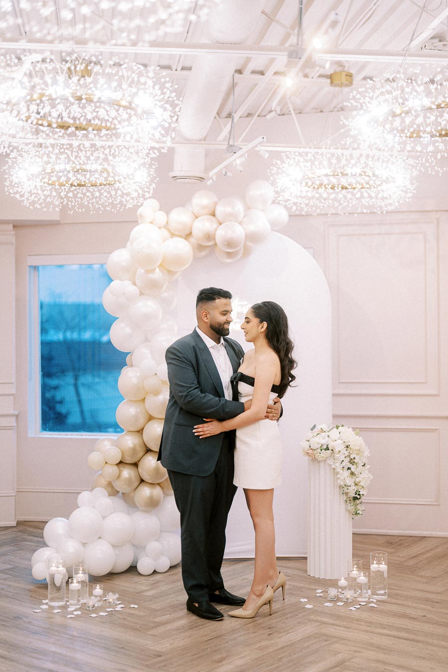 A couple embraces in a beautifully decorated room with white and gold balloons, elegant chandeliers, and floral arrangements, creating a romantic and sophisticated ambiance.
