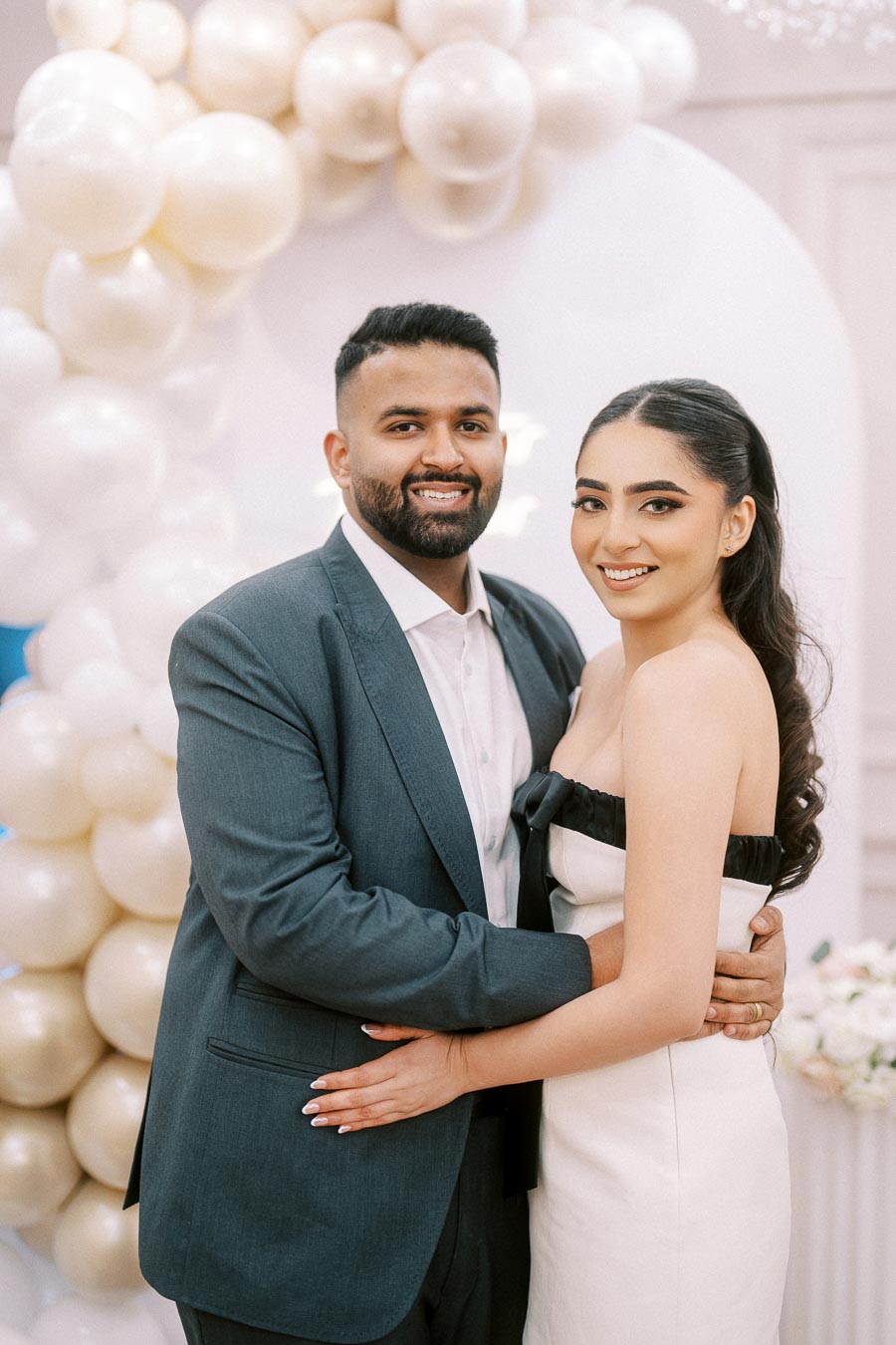 Smiling couple in elegant attire embracing in front of decorative white balloon arch at a celebration event.
