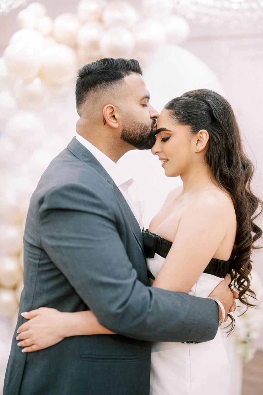 A couple embracing tenderly during an intimate moment at their wedding, with the groom gently kissing the bride’s forehead in a softly lit setting, surrounded by elegant decorations.
