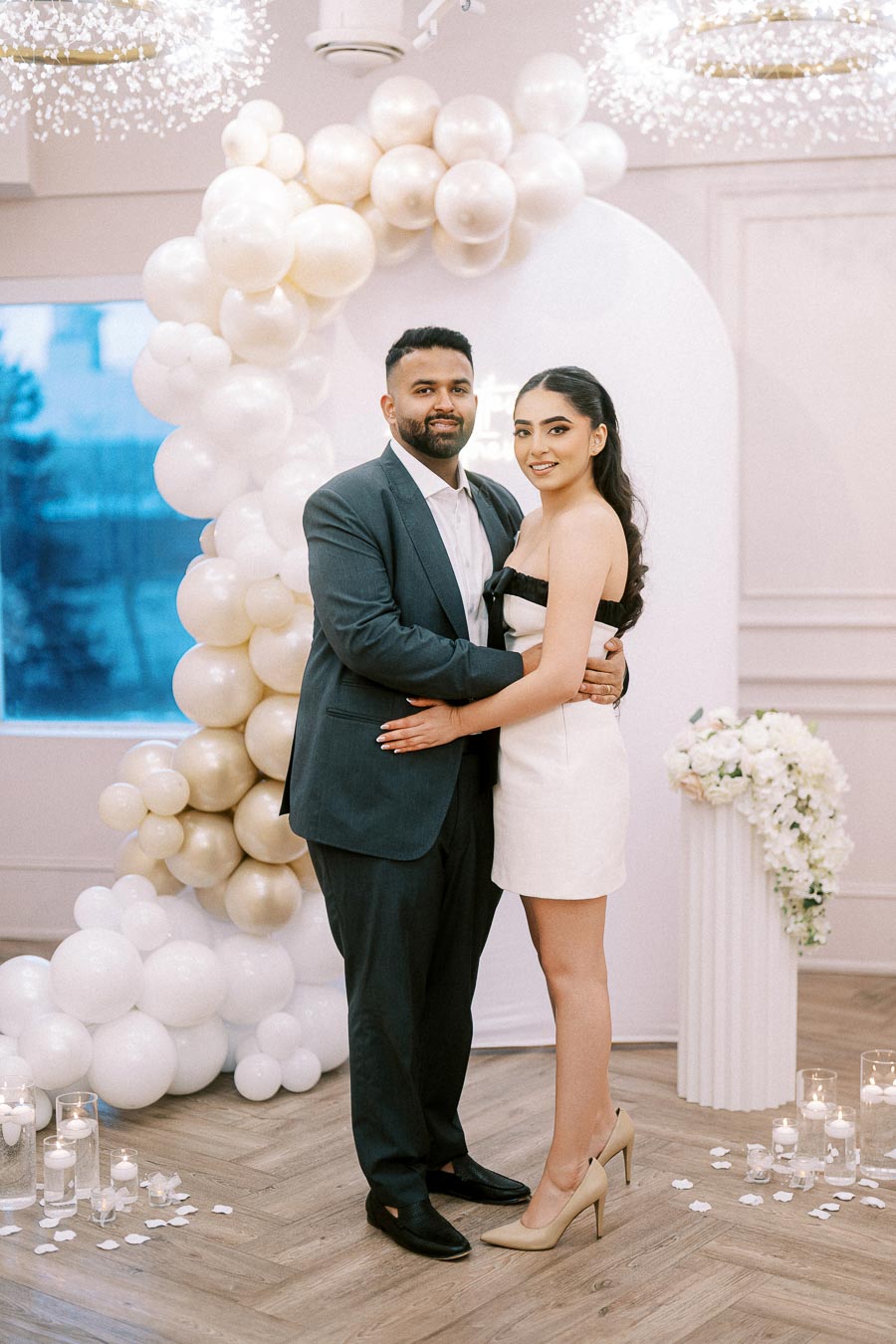 Elegant couple posing in a modern engagement setting with white balloon arch, candles, and floral decor.