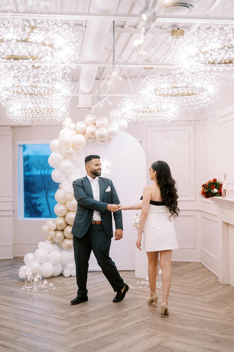 A couple dressed elegantly in formal attire shakes hands in a stylishly decorated venue featuring a balloon arch and ambient lighting, creating a romantic atmosphere.