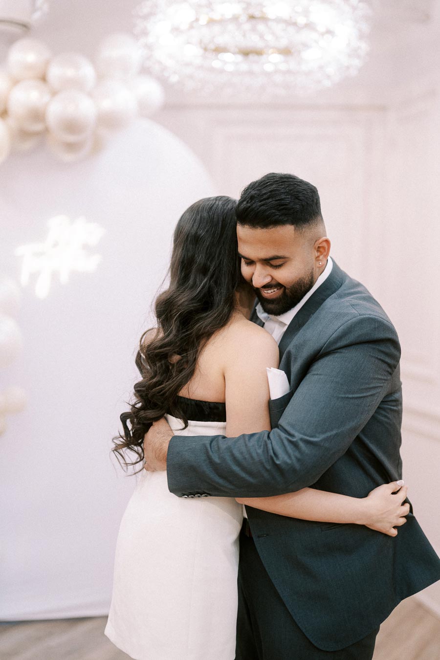 A couple embracing warmly during an elegant event, with the man wearing a dark suit and the woman in a white dress. The backdrop features soft lighting and decorative balloons, creating a romantic atmosphere.