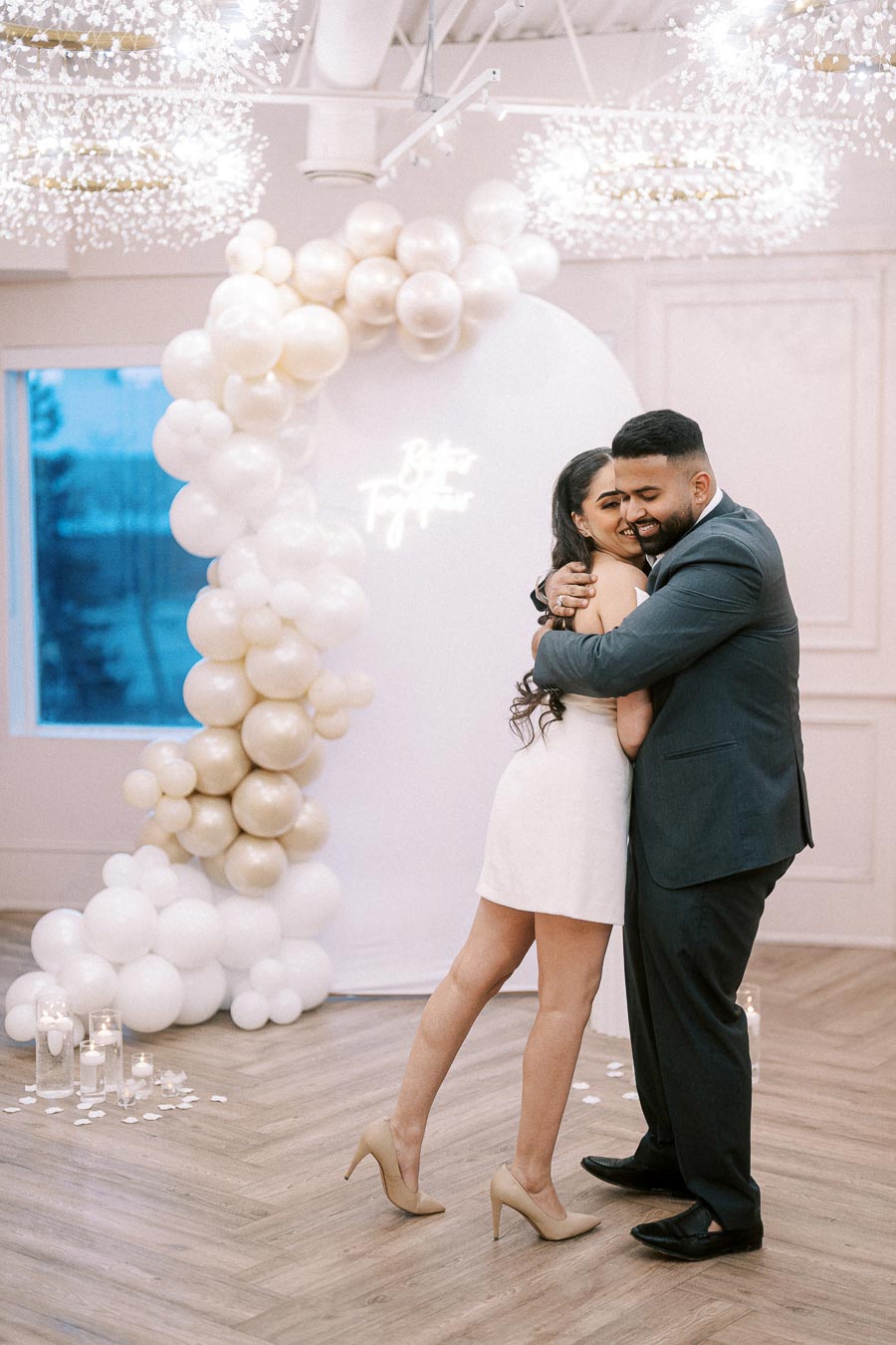A couple happily embraces in an elegantly decorated venue, featuring a backdrop of white and champagne balloons with a Better Together sign.