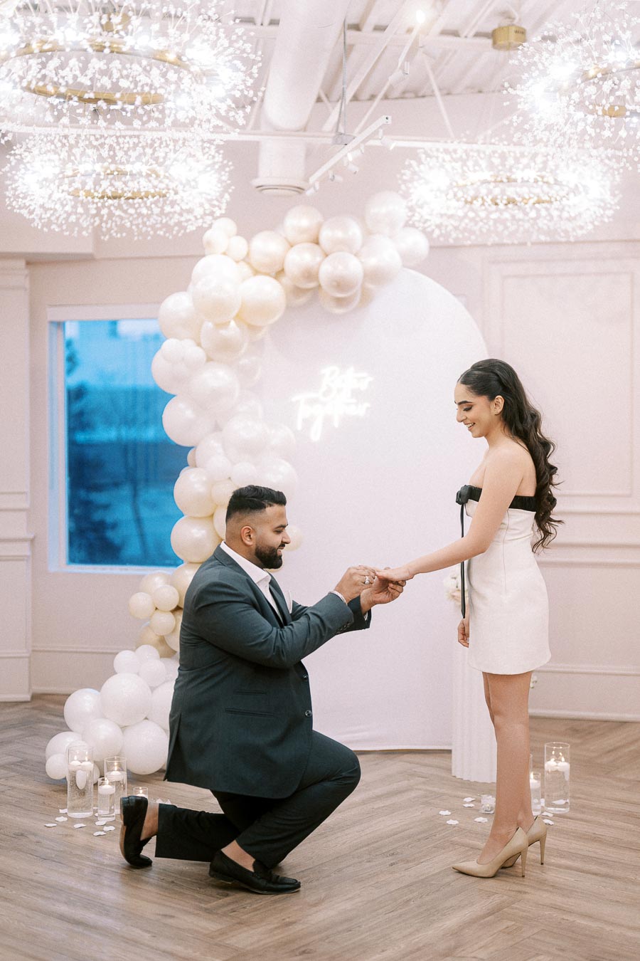 A man in a suit kneels and places a ring on a woman's hand in an elegant setting with white balloon decorations and soft ambient lighting, symbolizing an engagement or proposal celebration.