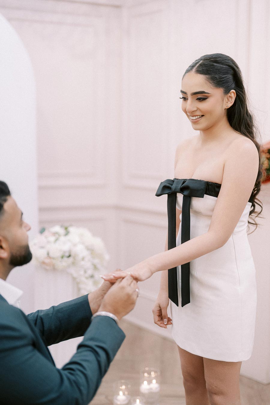 Man proposing to a woman in an elegant white dress with a black bow, set in a beautifully decorated room.