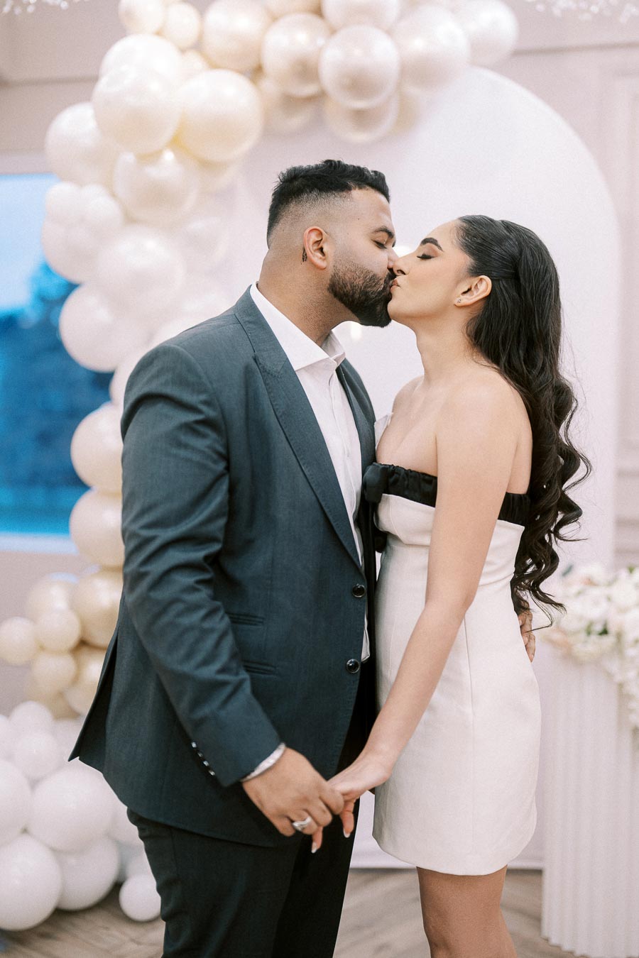 A couple shares a romantic kiss at an elegantly decorated wedding venue, surrounded by white balloons and floral arrangements. The man is dressed in a grey suit, while the woman wears a stylish strapless white dress.