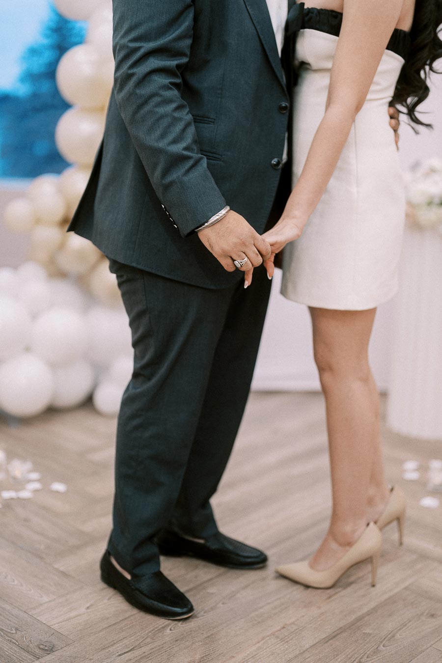 A couple holds hands during a formal celebration, showcasing their elegant attire with the man in a dark suit and the woman in a white dress and heels, surrounded by balloon decorations.