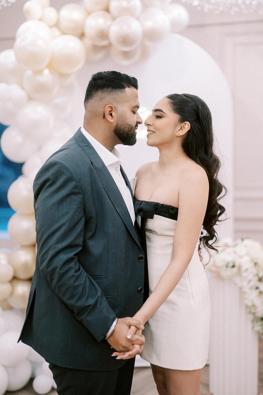 A couple embracing tenderly in front of a balloon arch decorated with cream-colored balloons, both dressed elegantly for a special occasion.