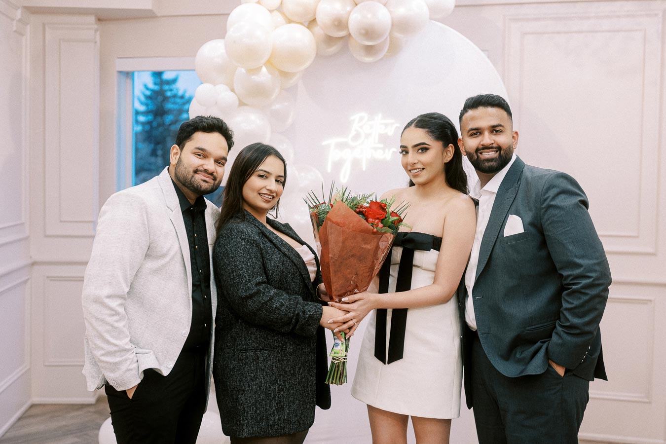 A group of four people smiling and posing together indoors, with one person holding a bouquet of red flowers. The background features a balloon arrangement and a sign that reads Better Together, capturing the essence of celebration and togetherness.