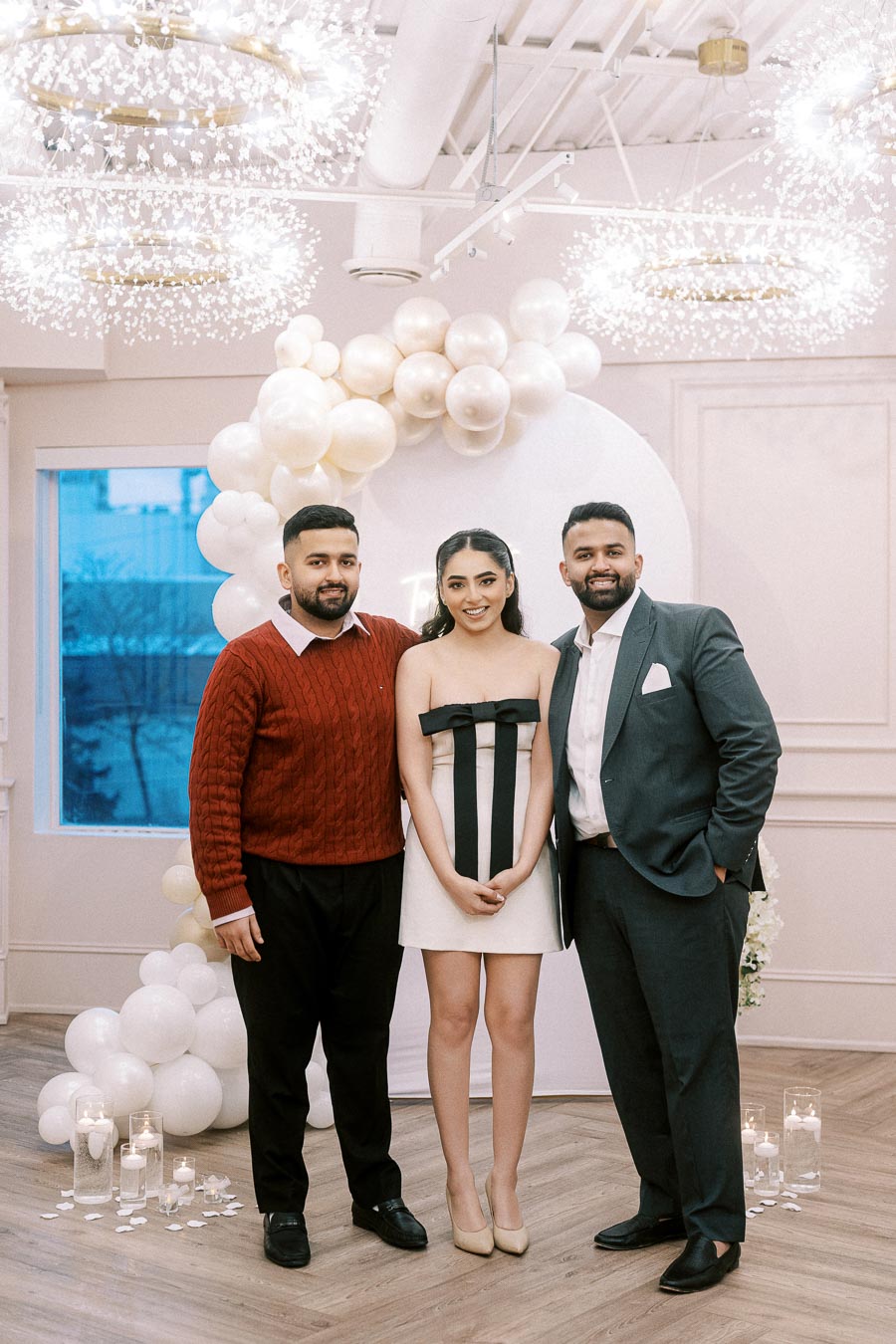 Three people posing together at an elegant indoor event, featuring a decorative balloon arch and warm lighting, creating a festive atmosphere.