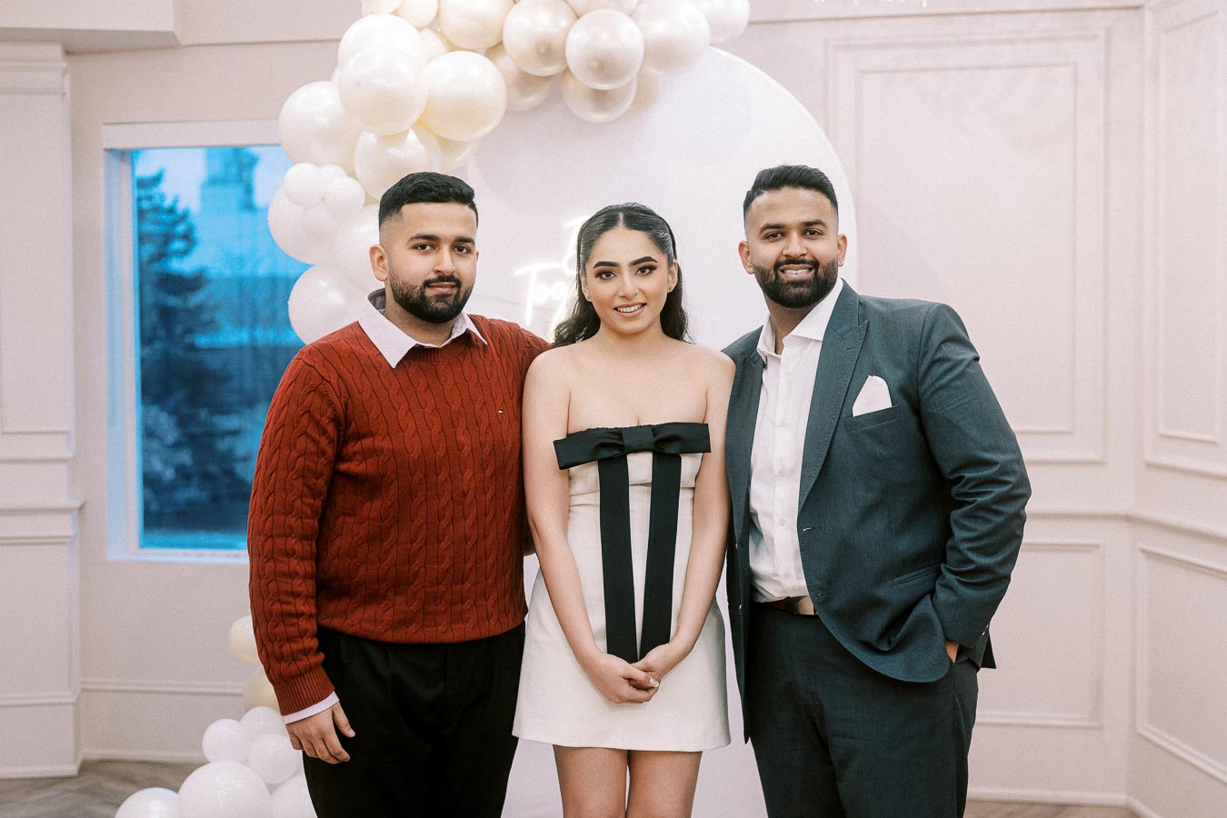 Group of three people standing together at an elegant event, with a white balloon arch in the background. The woman in the center wears a stylish white dress with a black bow, flanked by men in a red sweater and a blue suit.