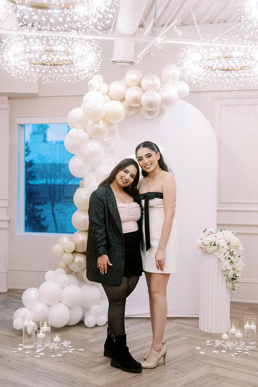 Two women smiling and posing at an elegant indoor event with a white balloon arch and floral arrangements. The setting includes modern lighting fixtures and decorative glass candles.