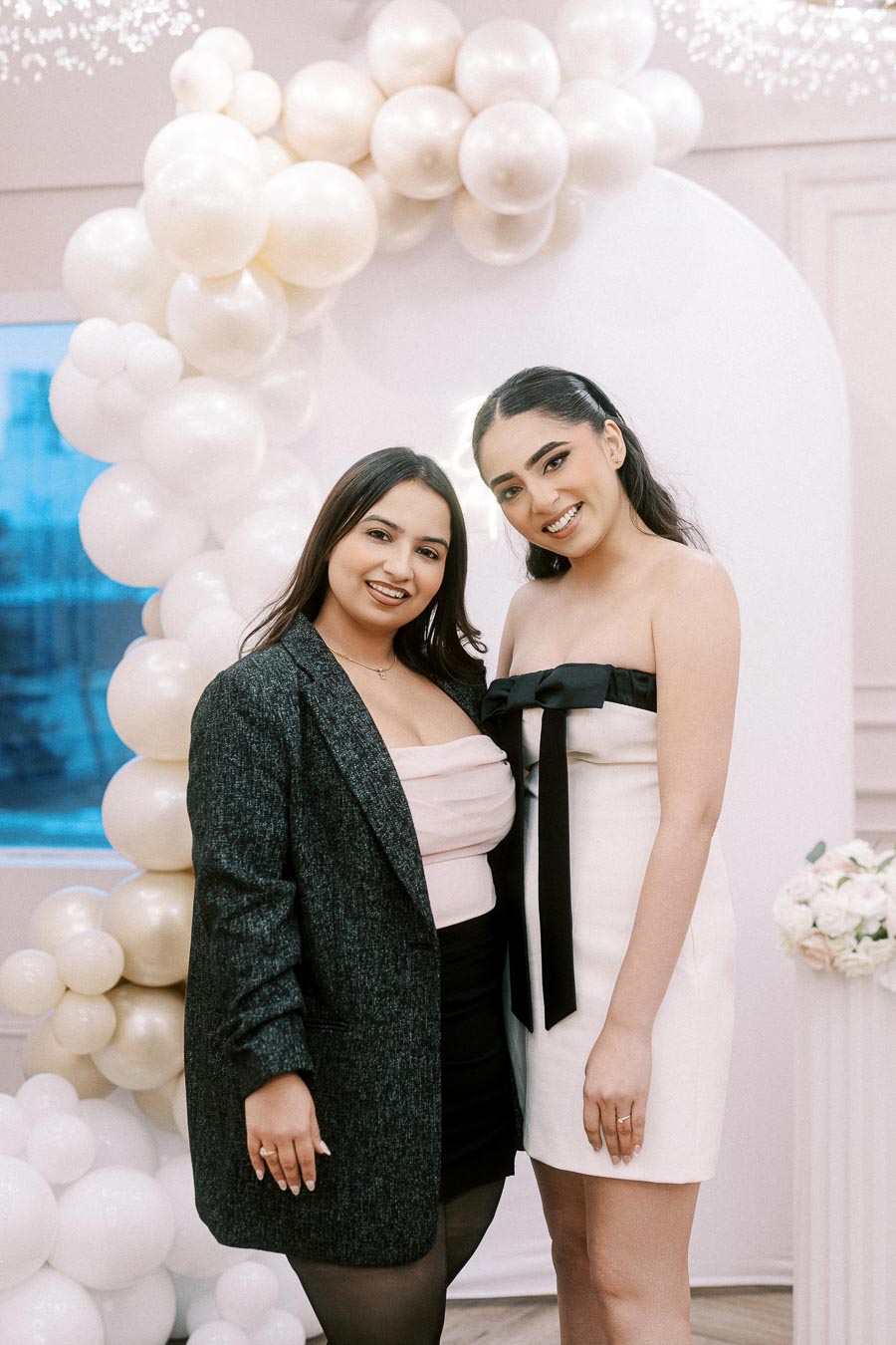 Two women smiling in front of an elegant balloon arch at a celebration event, with one wearing a grey blazer and the other in a white strapless dress with a black bow.