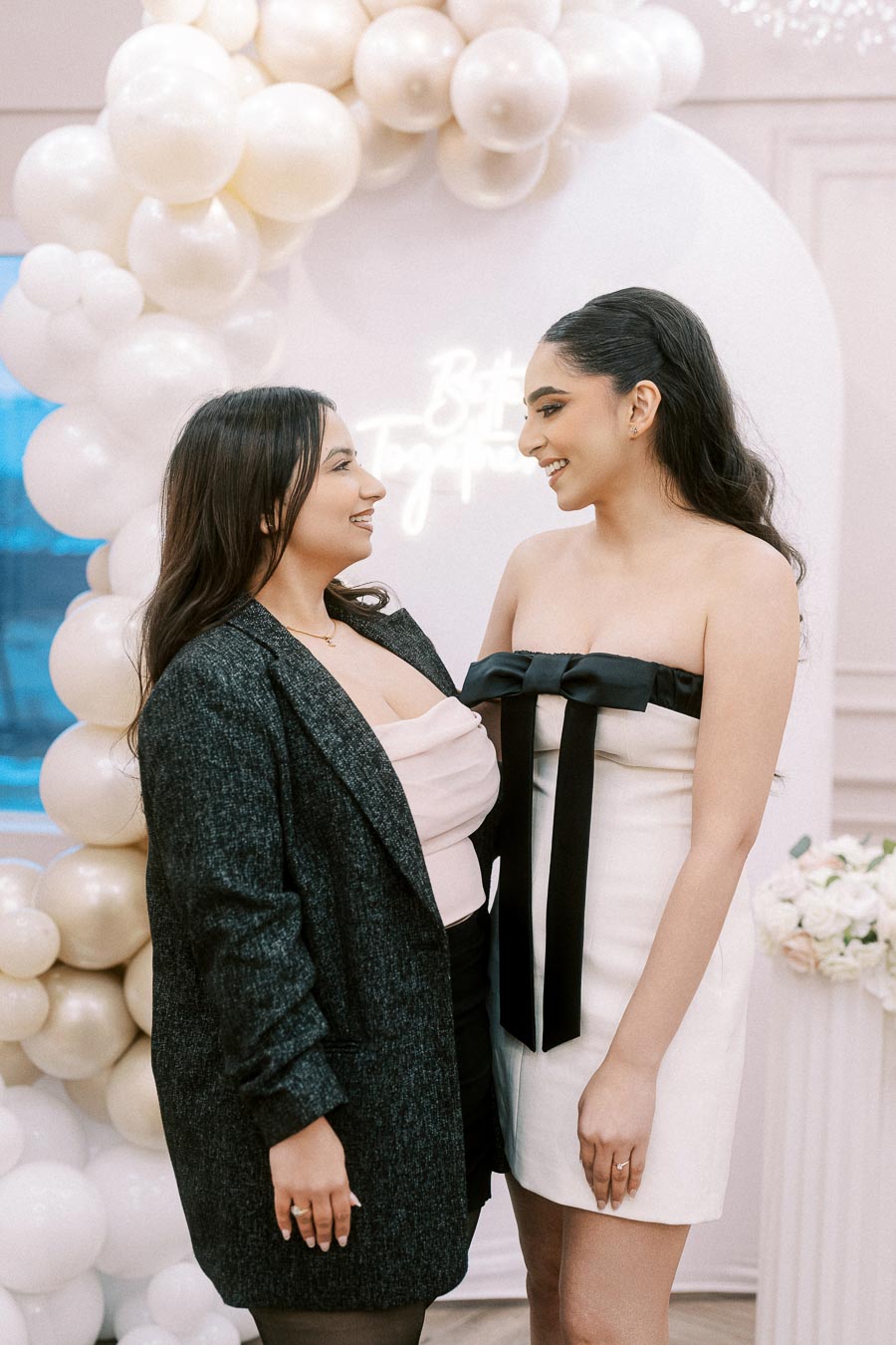 Two women smiling at each other in a stylish indoor setting, standing beside a decorative archway adorned with white balloons and flowers, exemplifying a festive or celebratory atmosphere.