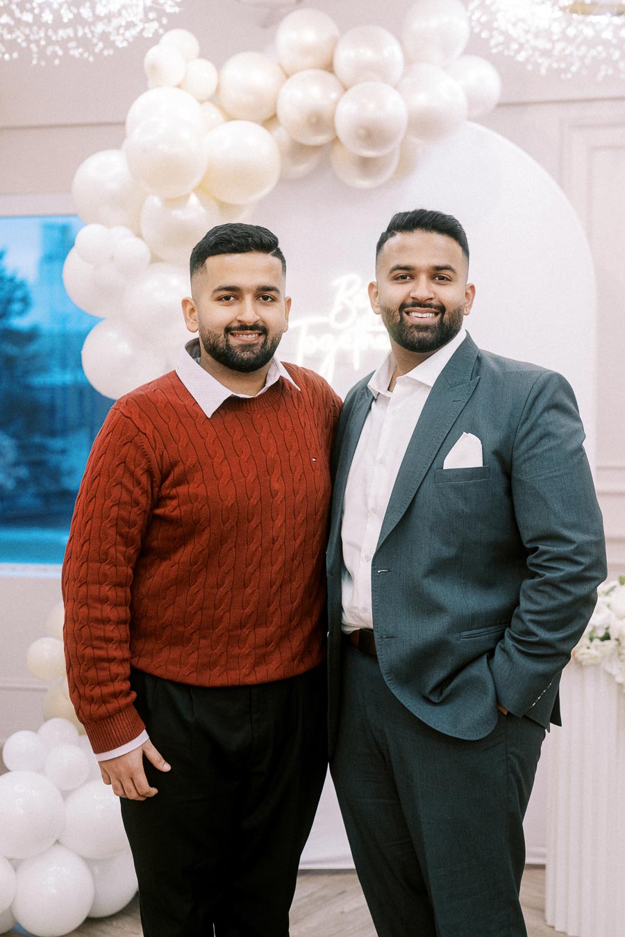 Two men posing together at an elegant event with white balloon arch backdrop, one wearing a red sweater and black pants, the other in a gray suit with a white shirt.