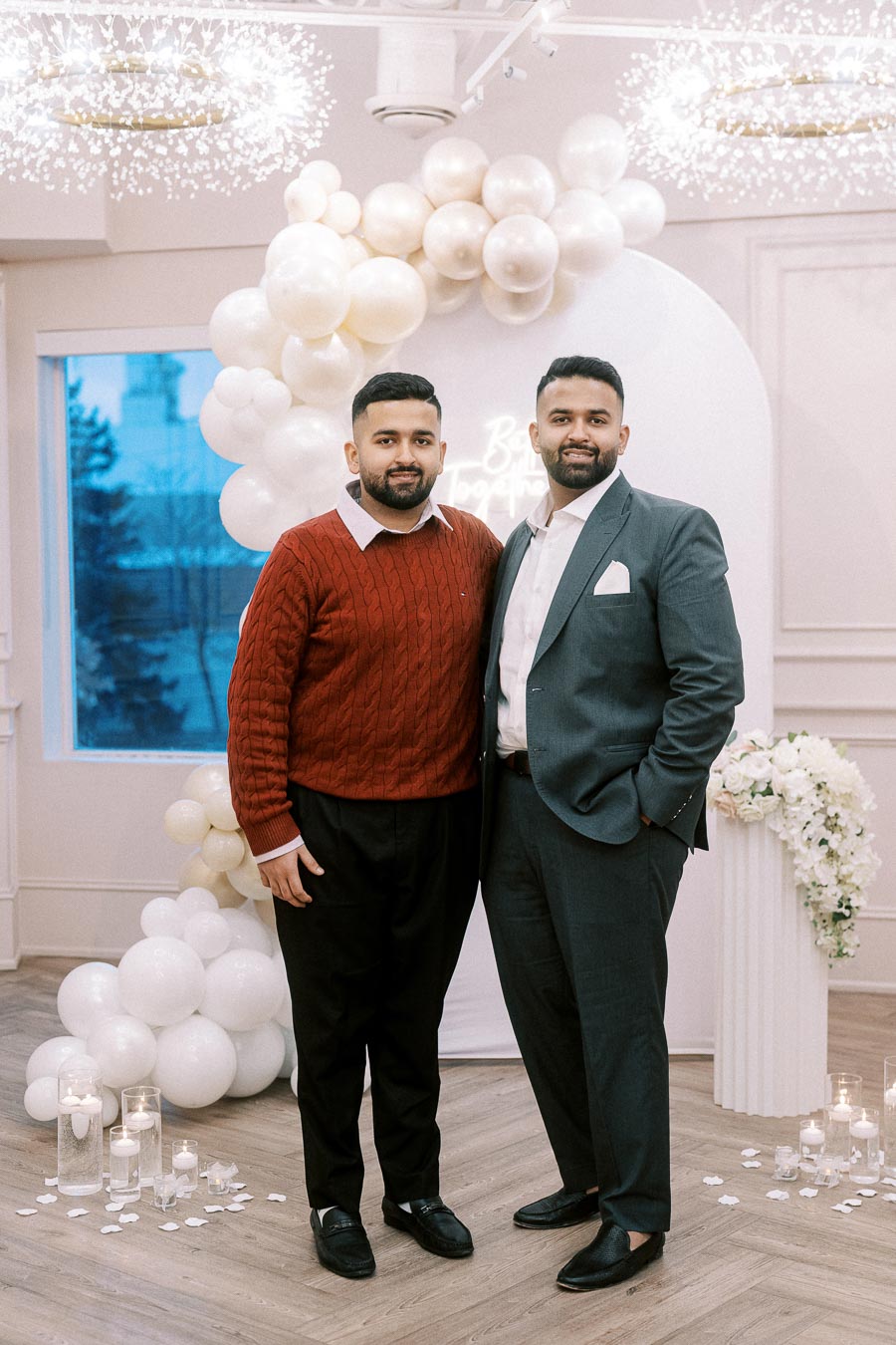 Two men dressed in formal and semi-formal attire stand in a decorated event space with white balloon arrangements and candle decorations, showcasing a stylish and elegant indoor celebration.