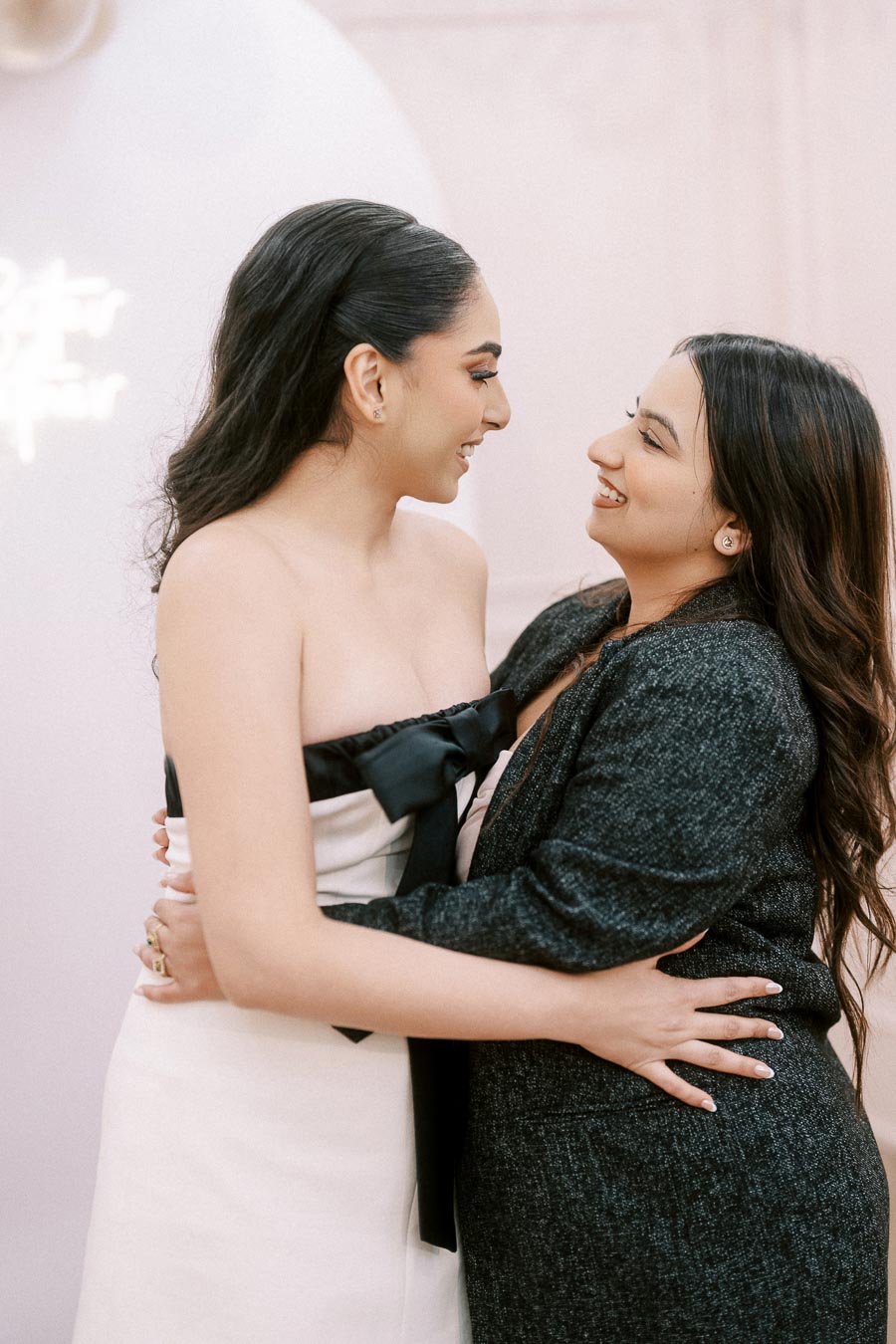 Two people warmly embracing and smiling at each other, with one wearing a strapless white dress with a black bow and the other in a black outfit, against a light-colored, softly blurred background.