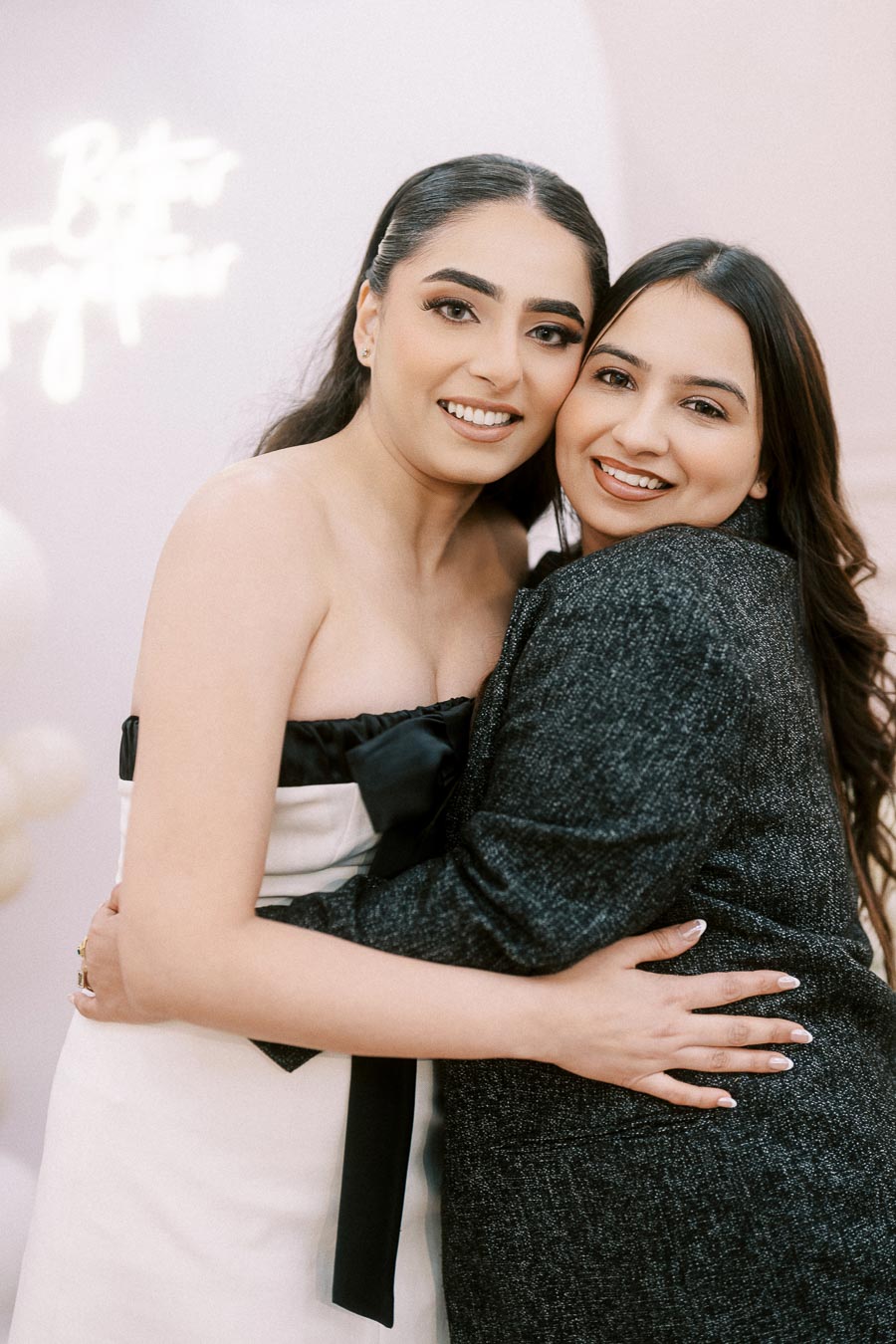 Two women smiling and embracing at an event with a soft, elegant background.