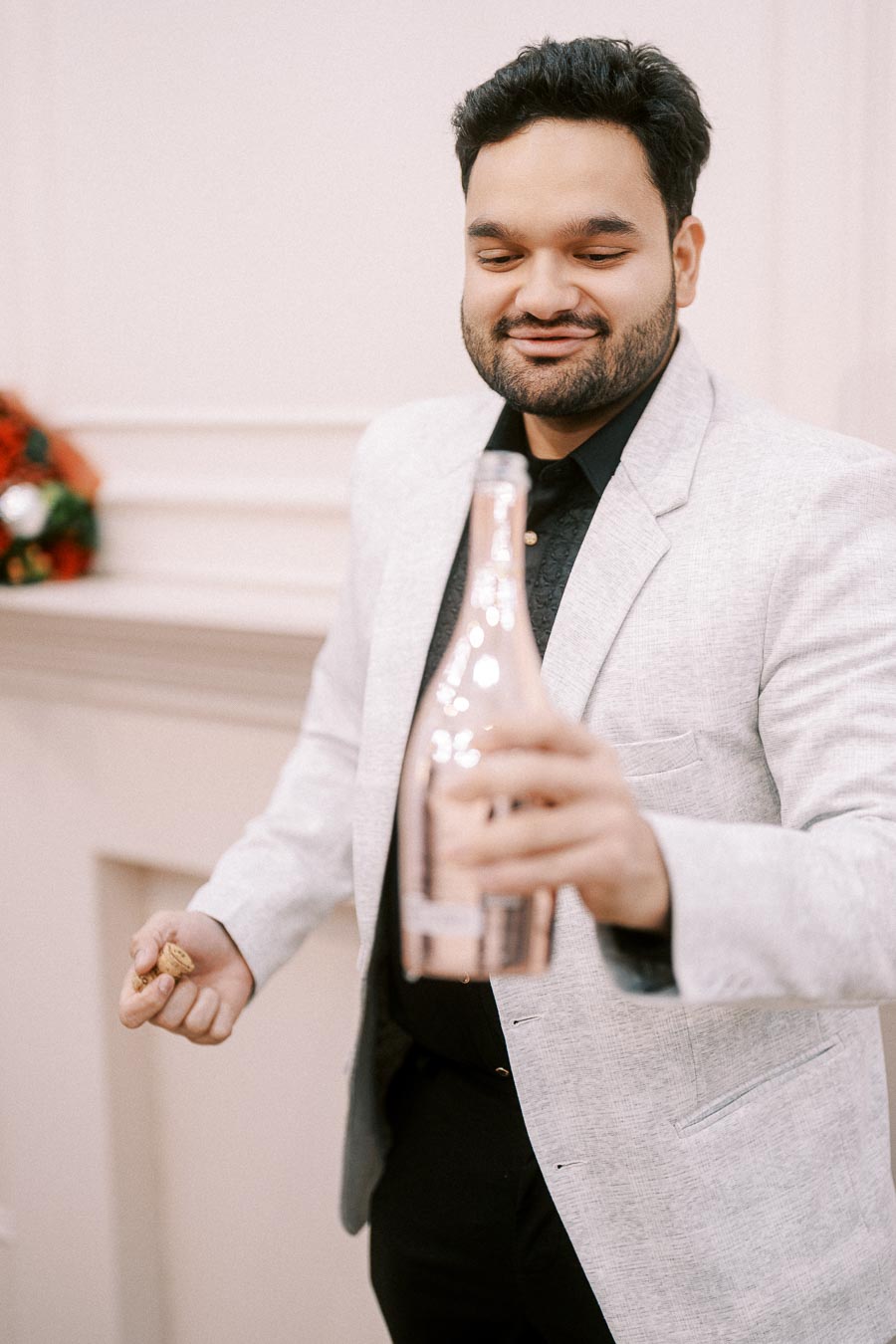 Man in a light gray suit opening a champagne bottle indoors, holding the cork, wearing a black shirt, celebrating a special occasion.