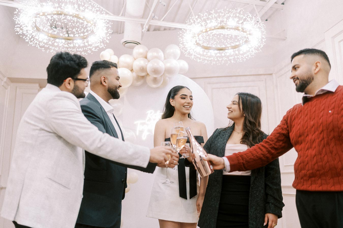 A group of five people celebrating with champagne glasses under elegant chandeliers and balloon decorations, creating a festive and joyous atmosphere.