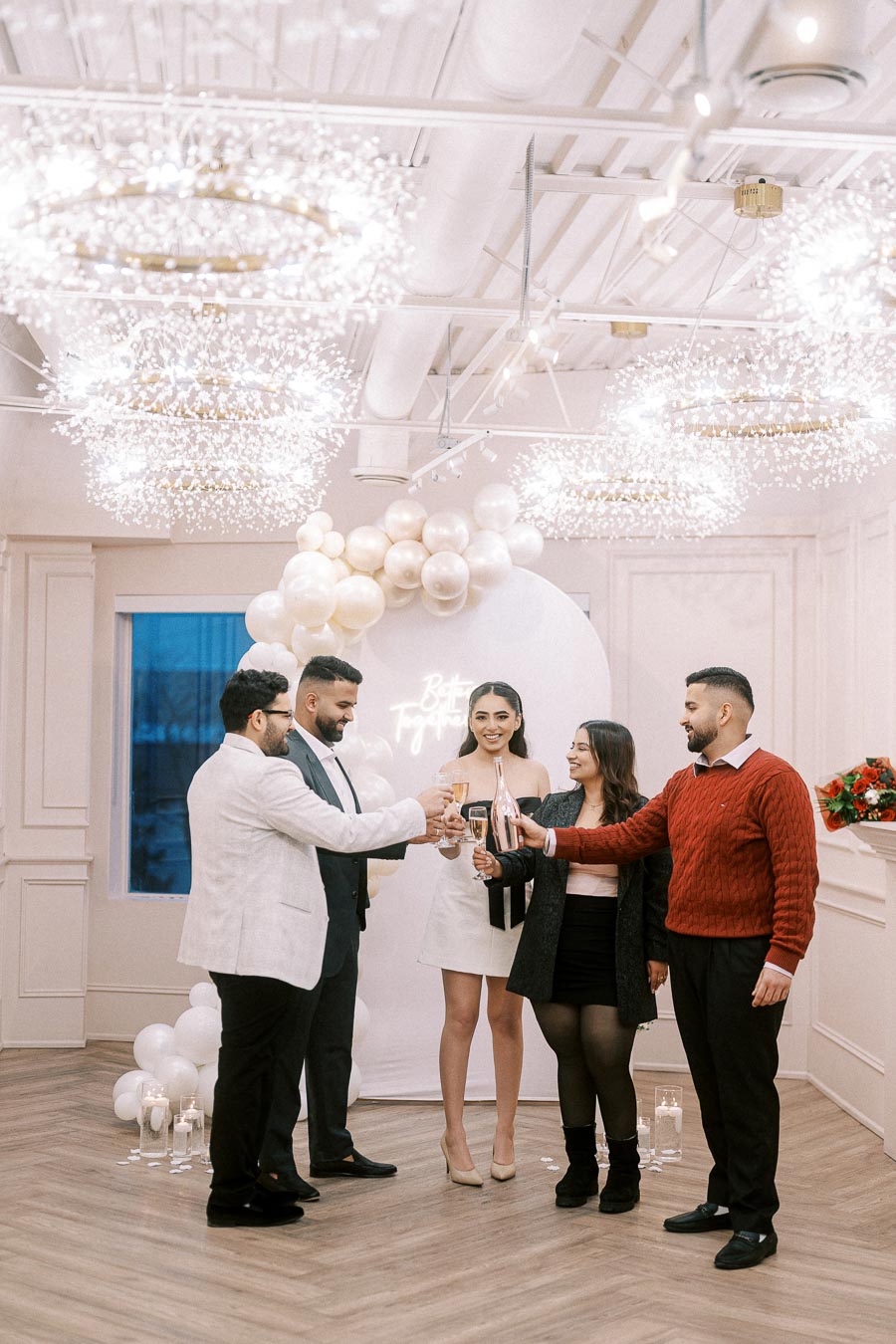 A group of elegantly dressed people toasting with champagne in a beautifully decorated room for a celebration, featuring white and gold balloons and sparkling chandeliers.