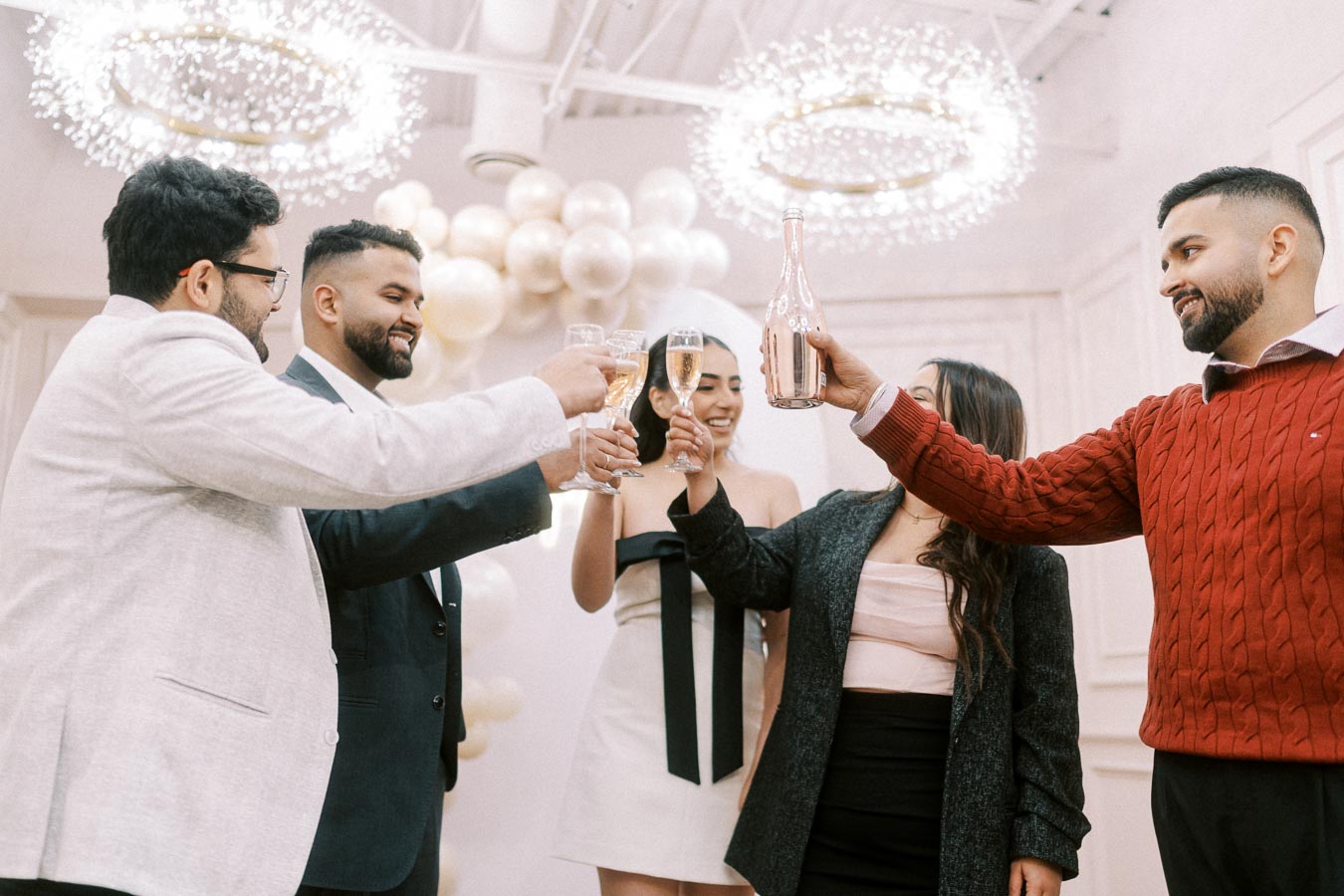 A group of five people celebrating with drinks at a festive indoor gathering, surrounded by elegant decor and balloon arrangements.