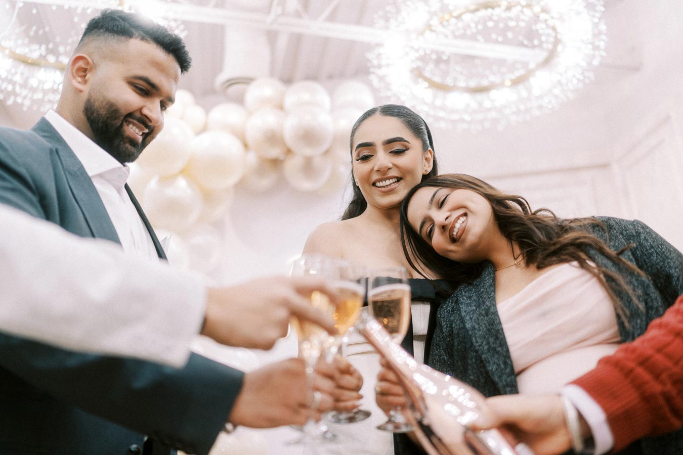 A group of friends celebrating with champagne glasses in a festive setting with decorative balloons and chandeliers.