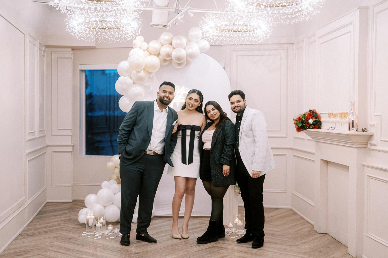 A group of people smiling and posing indoors at a celebration, with elegant white and gold balloon decorations and candles in the background, creating a festive atmosphere.