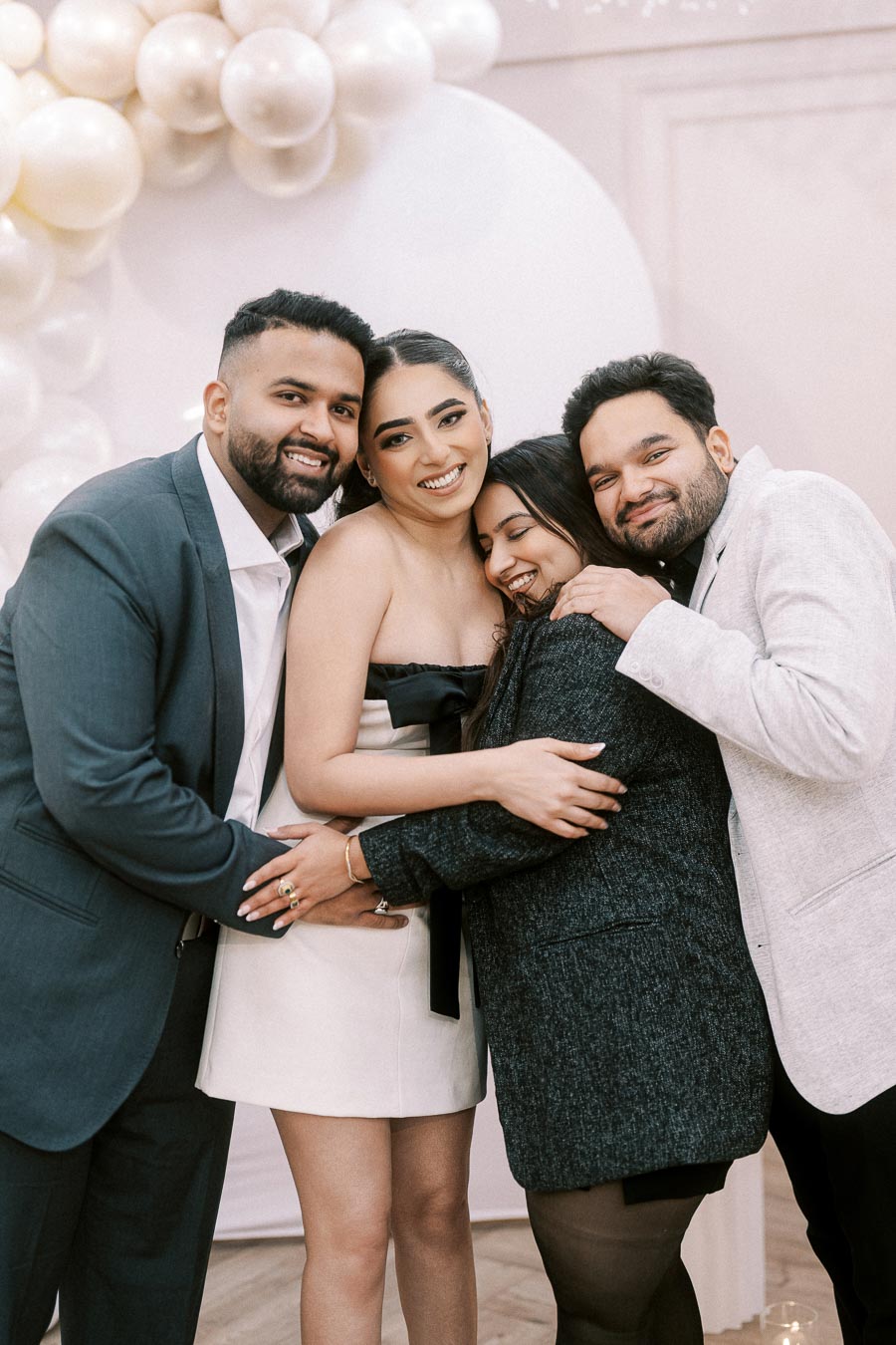 Group of friends hugging and smiling at a celebration event with balloon decorations in the background.