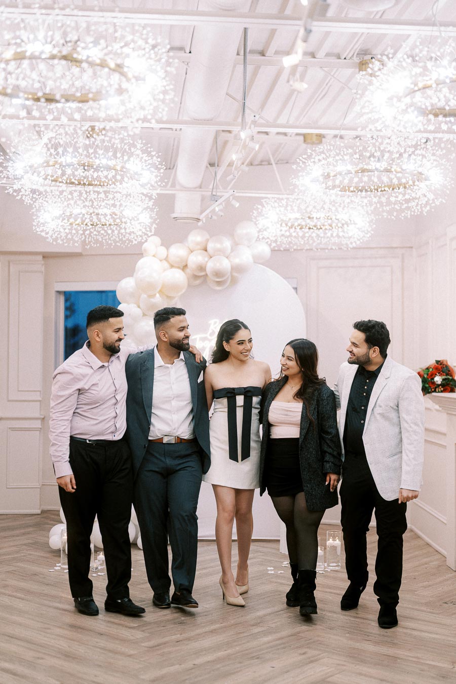 A group of five well-dressed people smiling and posing together at an elegant indoor event, featuring decorative lighting and white balloon arrangements in the background.