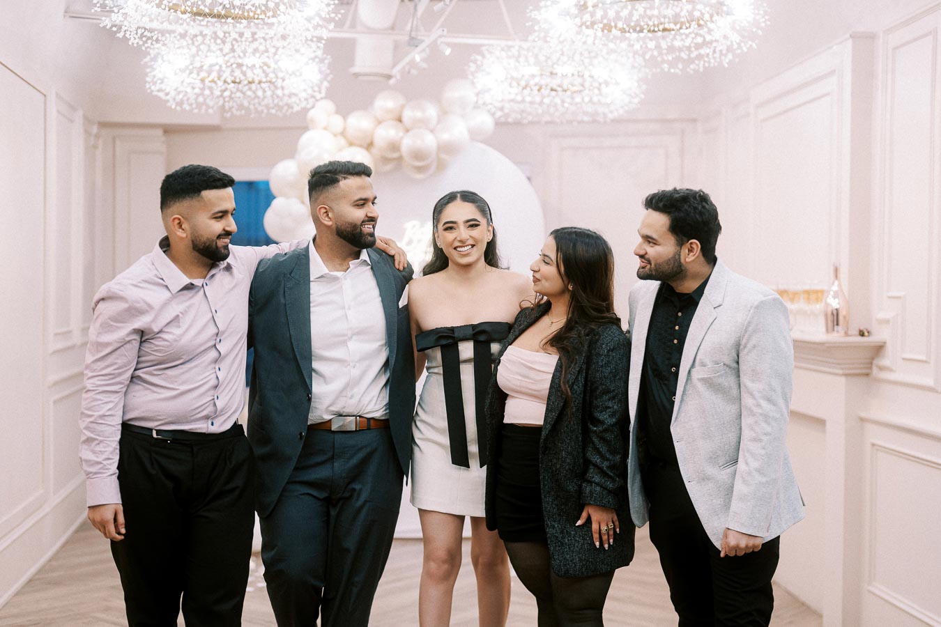 A group of five well-dressed people smiling and standing together at an indoor celebration with elegant decor, featuring balloons and chandeliers.