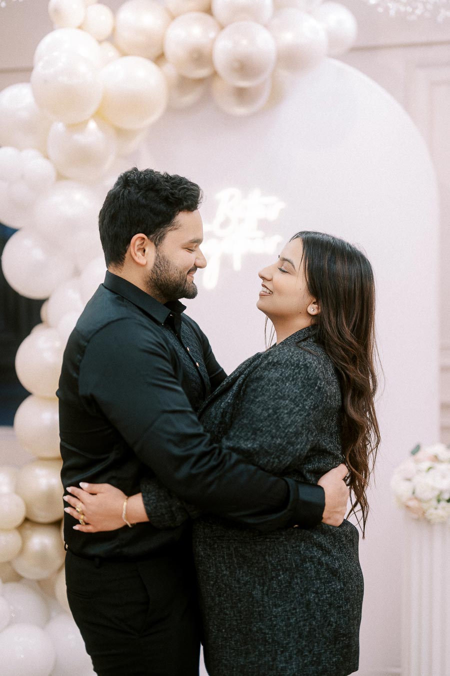 A couple embraces lovingly in front of a white balloon arch, smiling at each other, symbolizing romance and celebration.