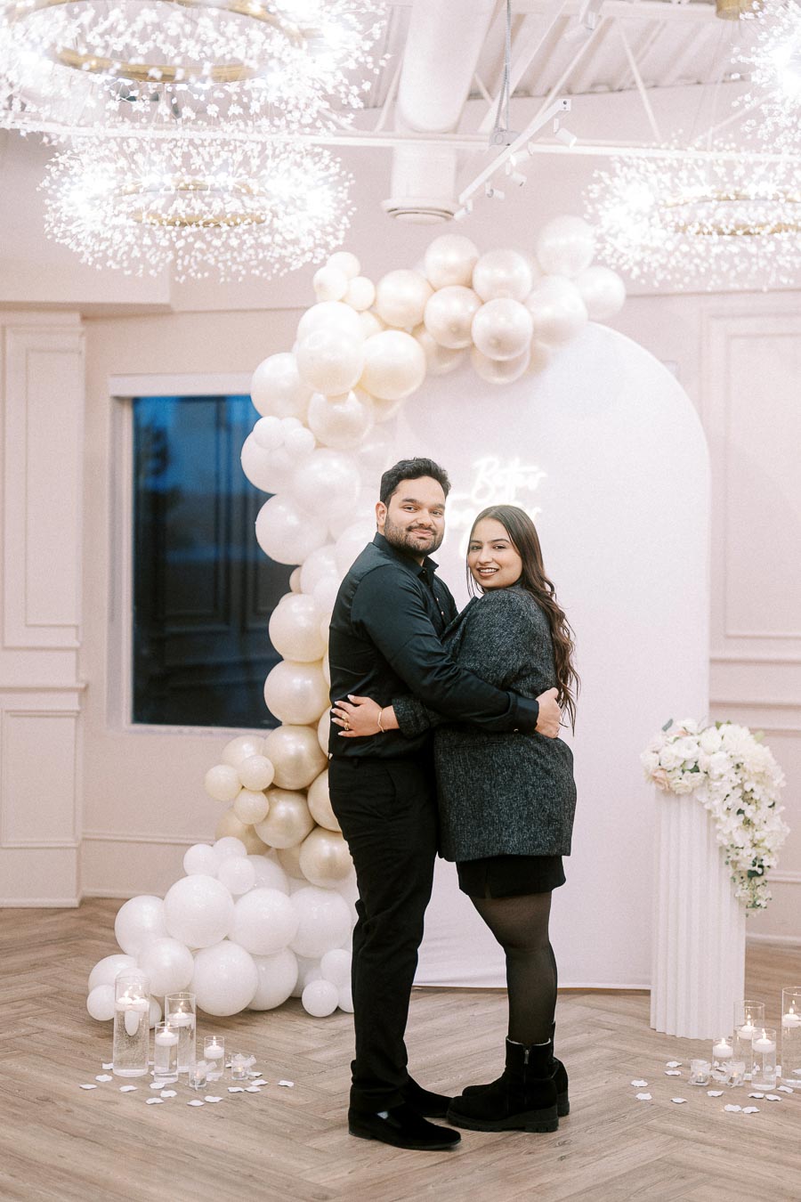 A couple posing together at an engagement or celebration event, surrounded by a decorative white and gold balloon arch, floral arrangements, and candles.