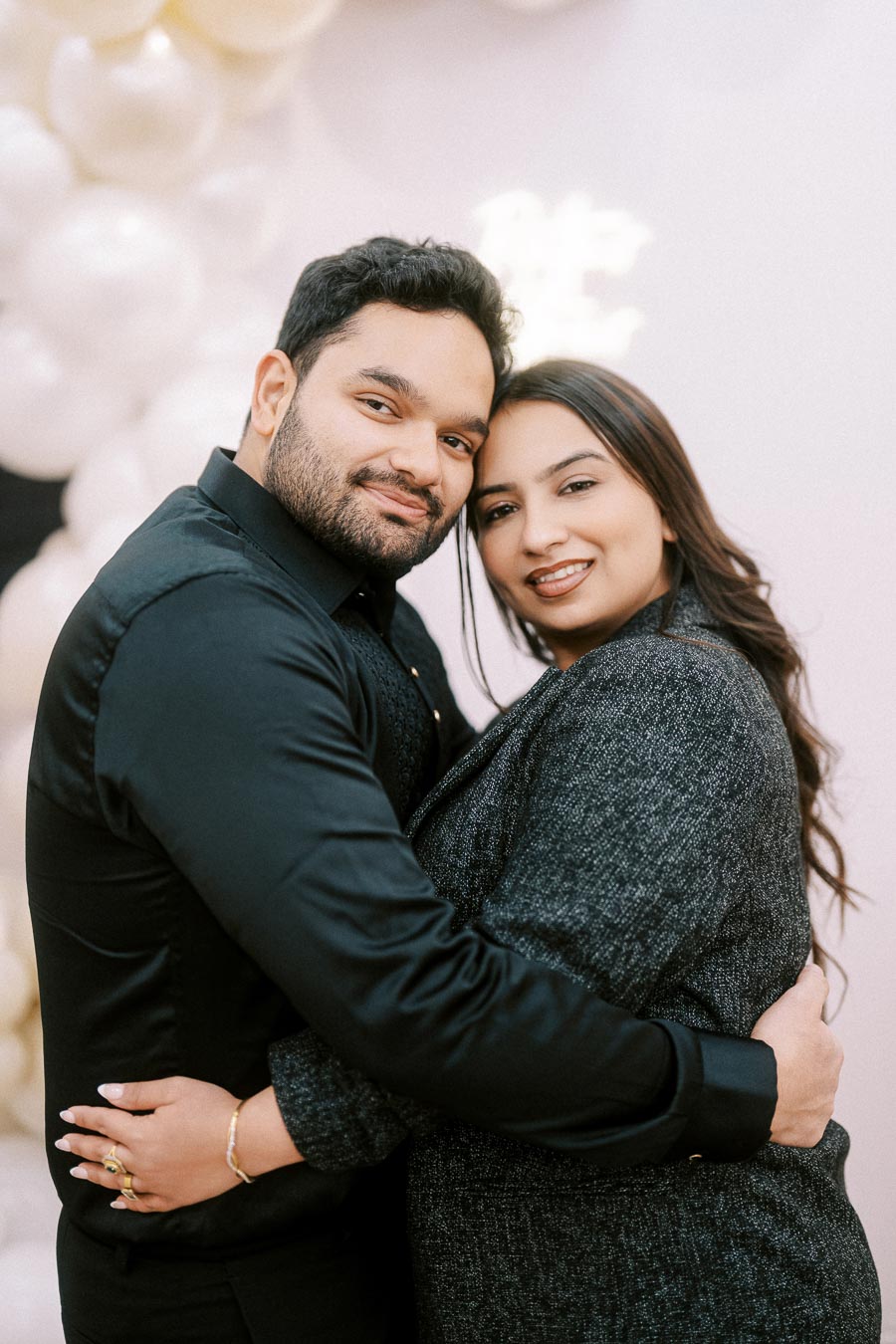 Happy couple embracing at a celebration with white balloons in the background, both smiling warmly and dressed in elegant dark attire.